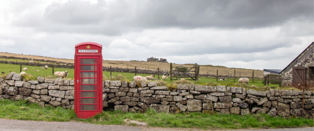 Photo of an old fashioned red phone box in front of a dry stone wall. Behind the wall is a field with sheep and a farm building.